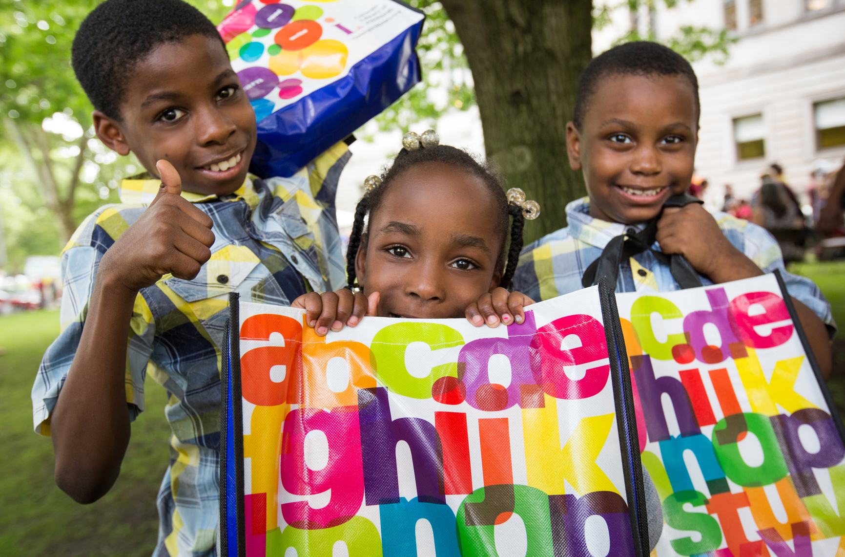 Three school children outside Carnegie Library of Pittsburgh, smiling and holding bags with colorful alphabet letters on them.