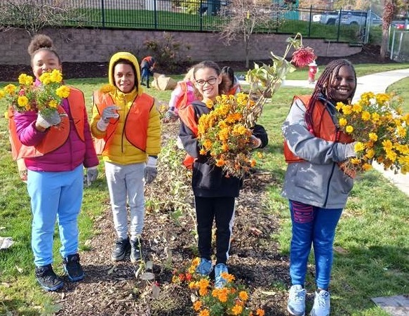 Four girls are smiling at the camera, holding flowers that they have removed from a garden. All four are wearing orange vests.