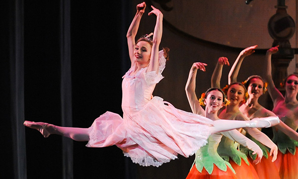 Ballerina wearing a pink dress leaping with arms overhead; a line of dancers standing and smiling behind her.