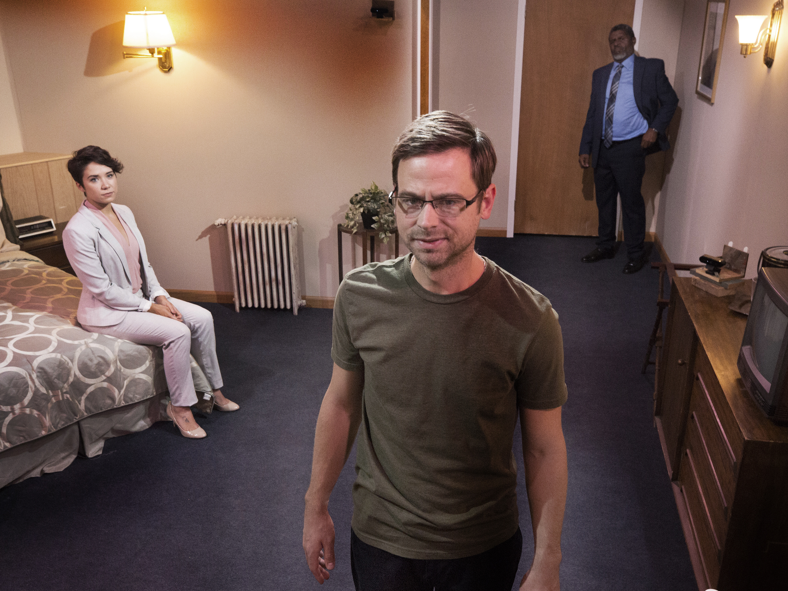 Three people in a hotel room; a woman sitting on the bed, a man standing in the foreground, and a man standing at the door.