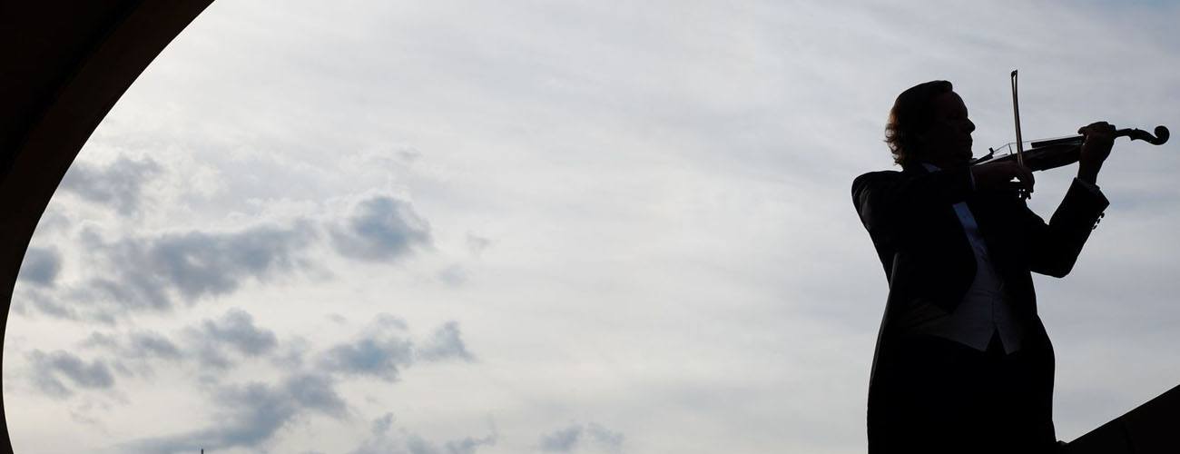 Silhouette of a man in a tuxedo playing a violin against the backdrop of a grey sky.