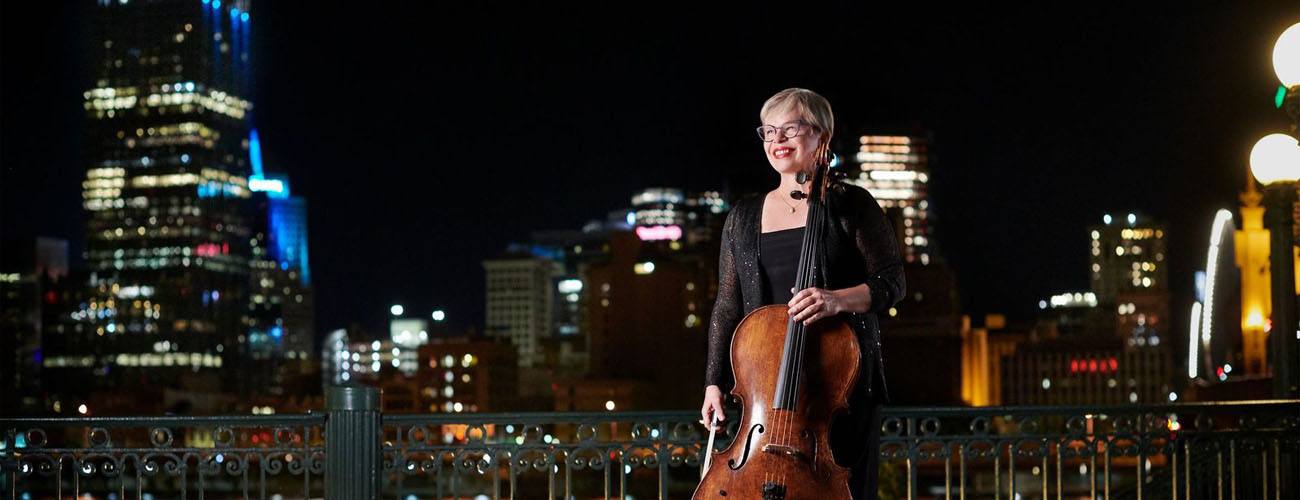 Woman in a black dress standing and smiling with a cello and the city at night in the background.