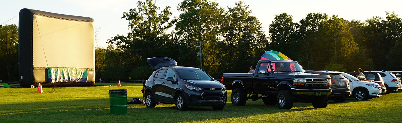 Cars and trucks parked on a large lawn at dusk in front of a large, inflatable movie screen.