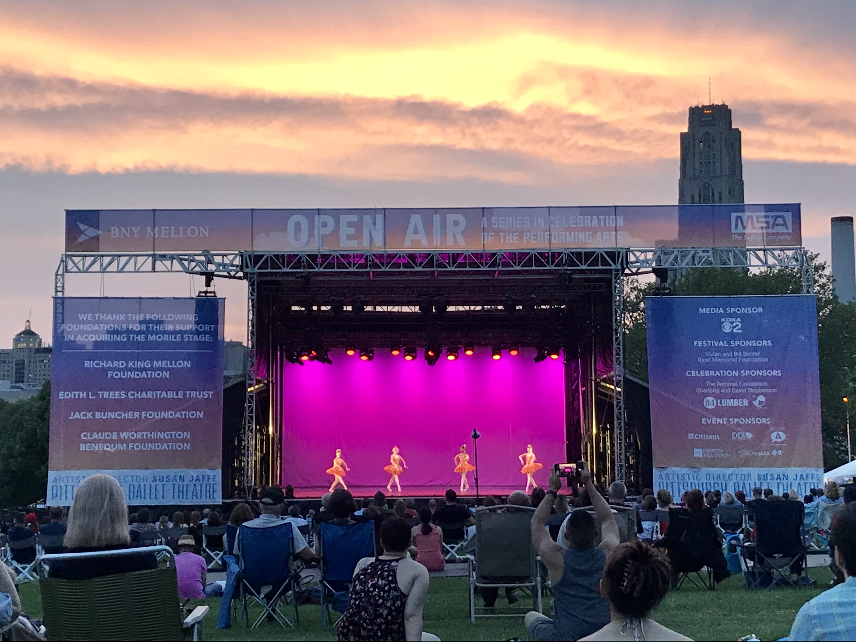 Pittsburgh Ballet Theatre's Open Air stage at dusk with pink lighting and four dancers onstage; audience watching from the lawn on Flagstaff Hill in Schenley Park.