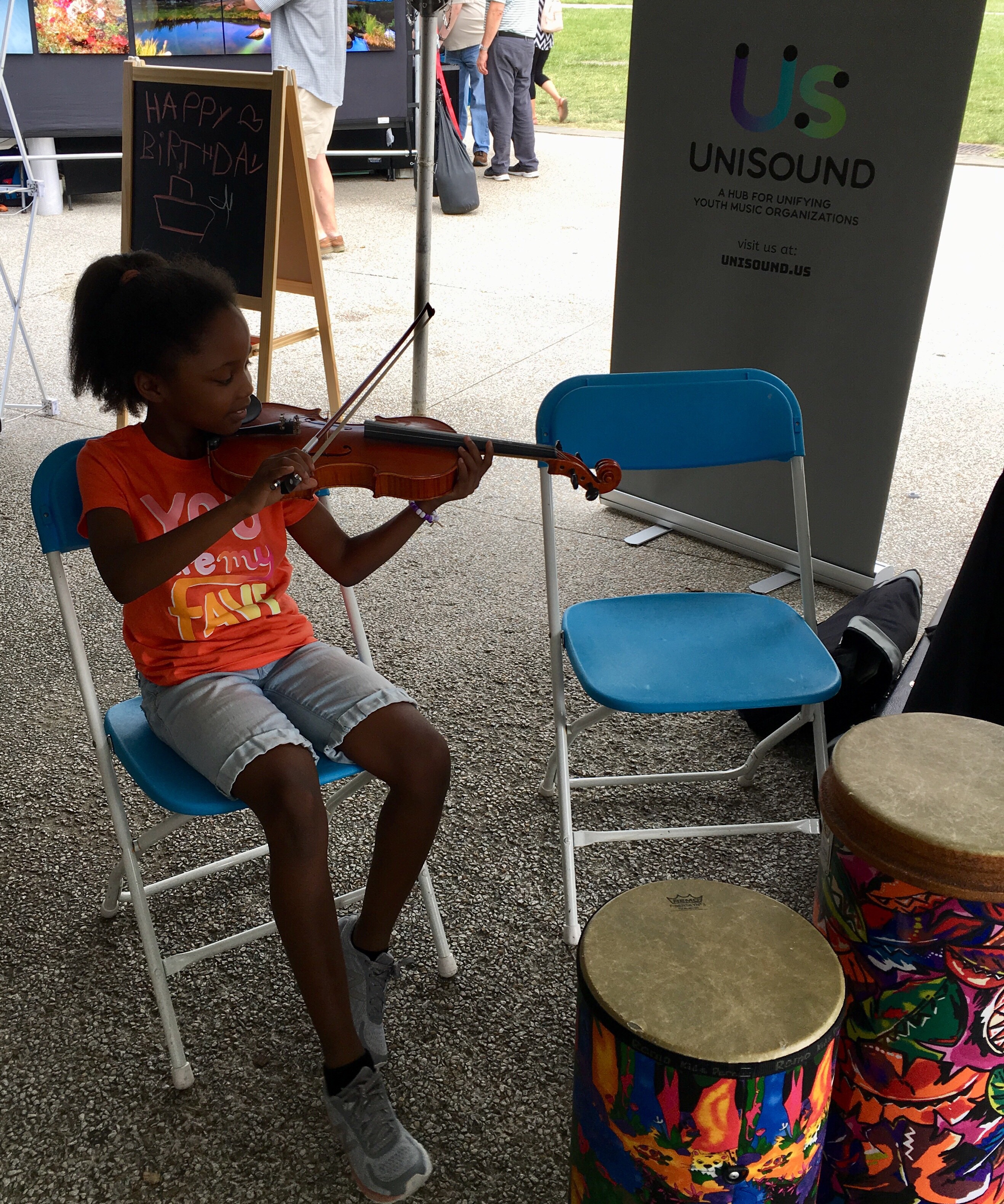 A young girl sits on a blue chair outside, smiling and playing the violin.