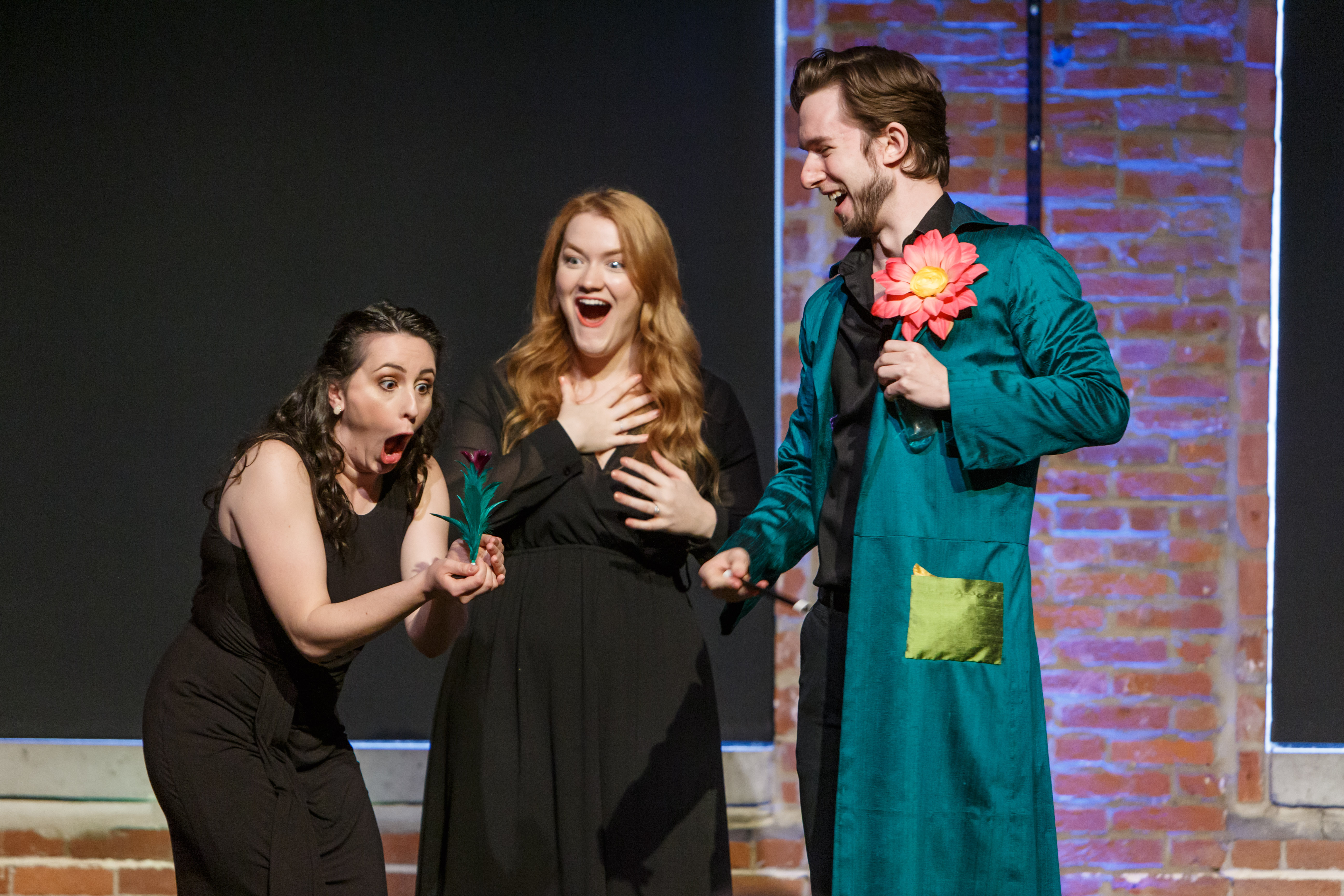 Three white performers on a stage, reacting with funny faces to the sight of a flower