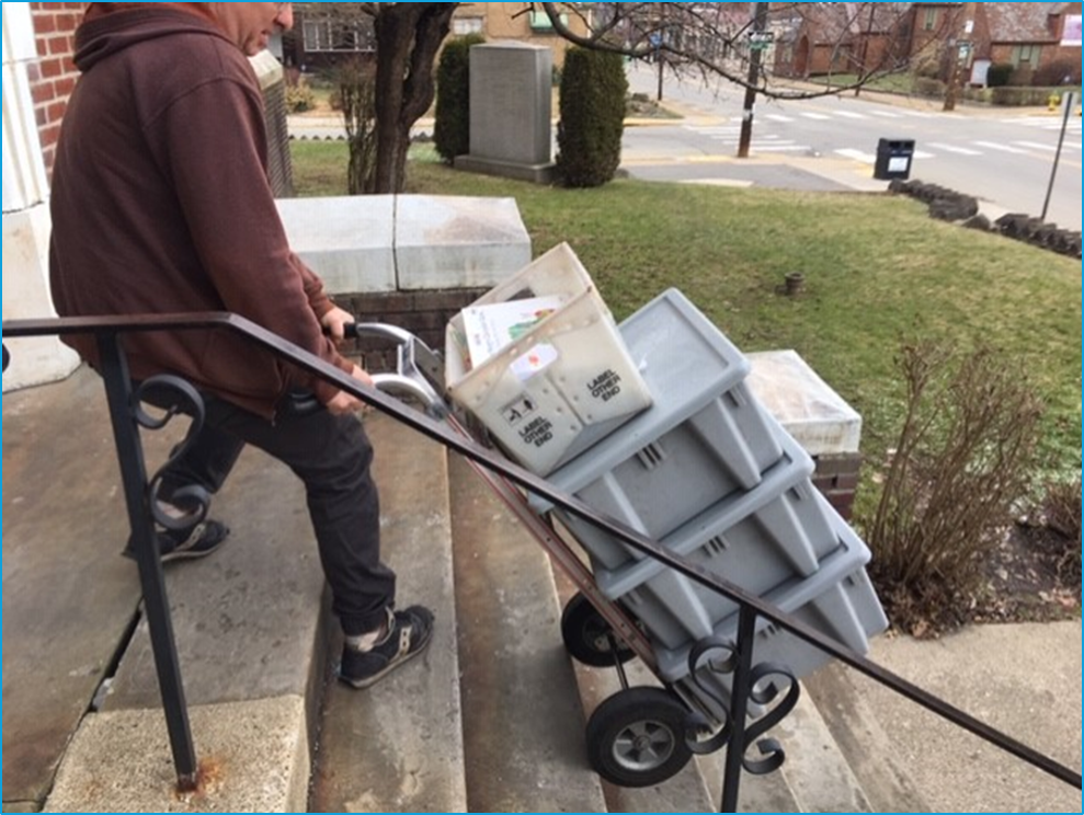 Shows a man using a hand cart to take boxes of library books up stairs