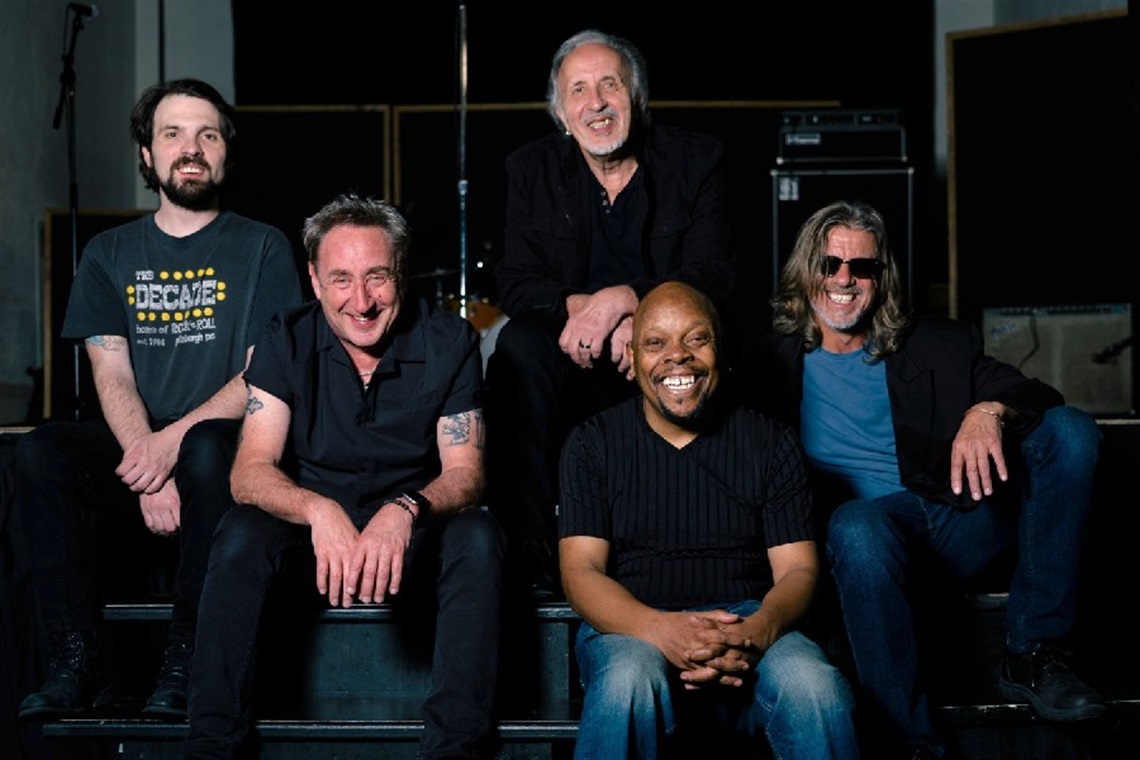 Photo of the five members of Joe Grushecky & The Houserockers sitting in front of a stage, smiling for the camera
