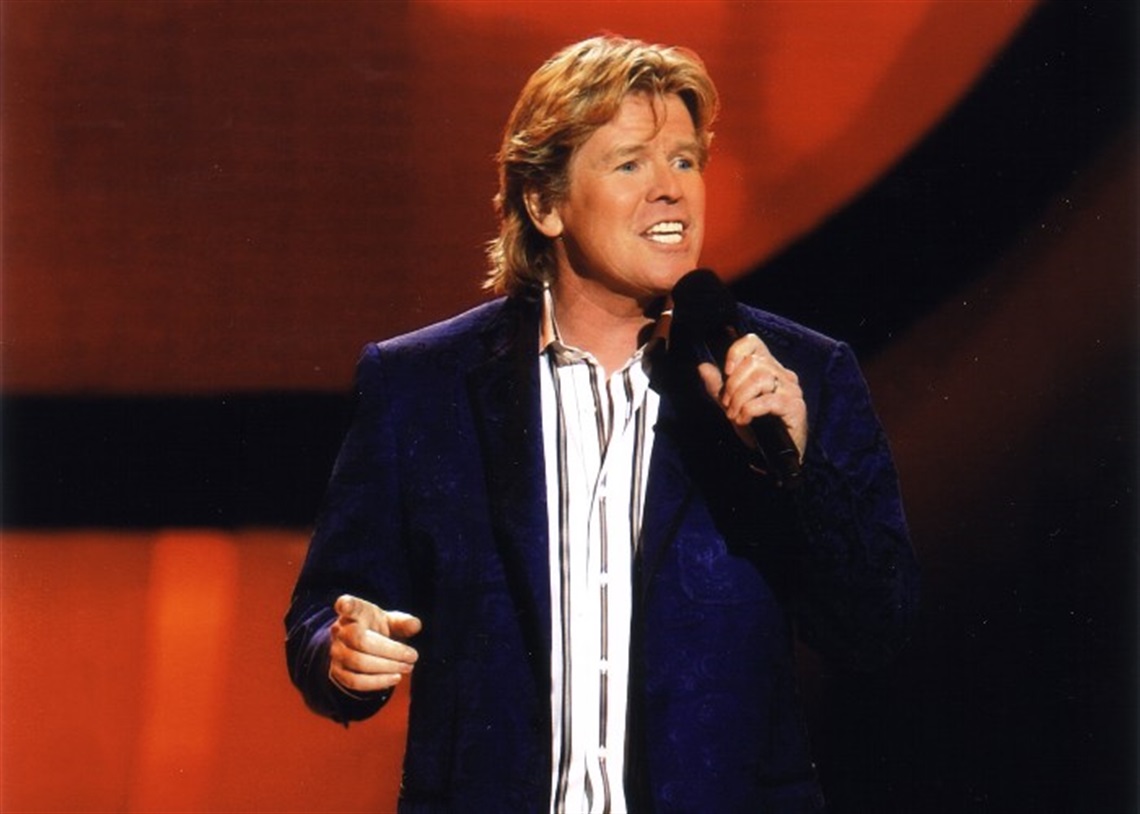 Photo of Peter Noone singing on stage in a dark purple coat