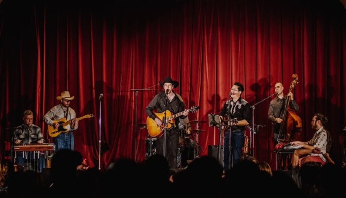 Photo of six performers on stage in front of a red curtain with country western outfits and instruments, including guitars, a bass and steel guitar