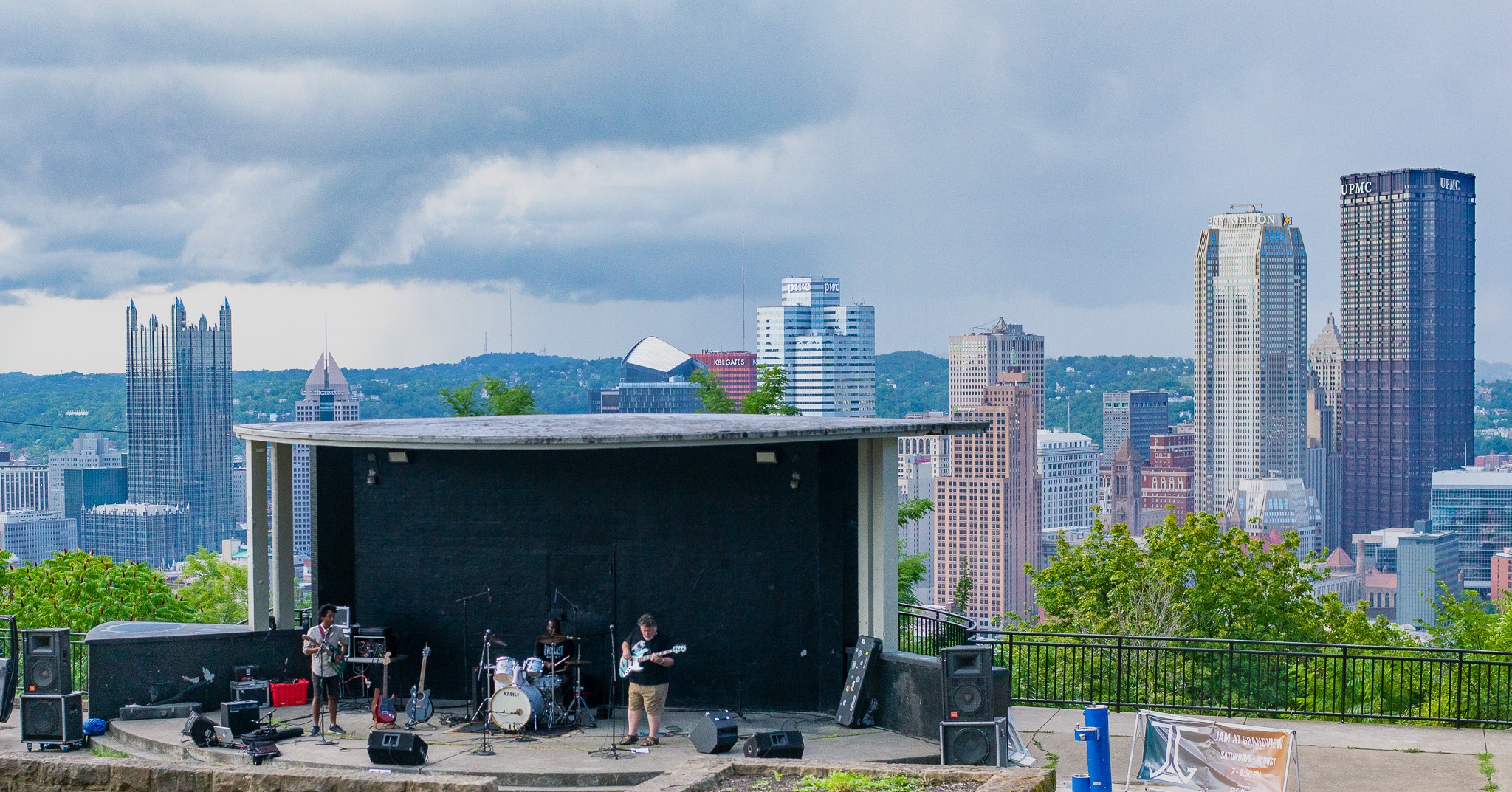 Photo shows a rock band playing at a bandshell in Mt. Washington with the Downtown Pittsburgh skyline behind it