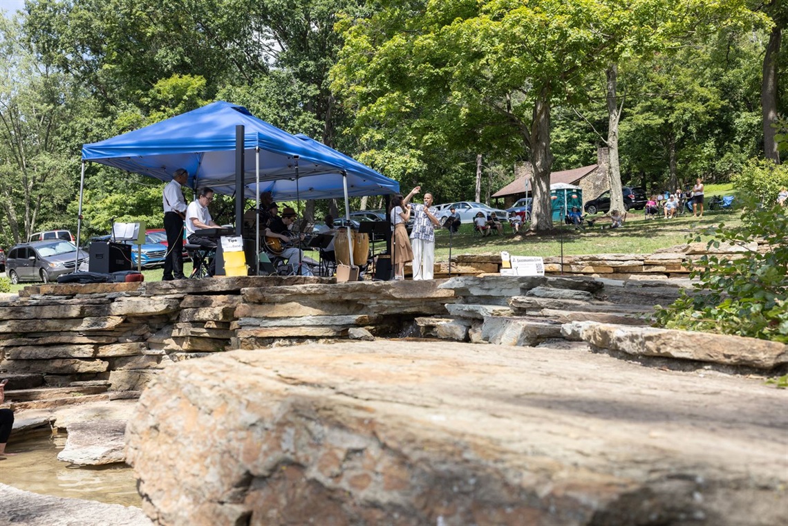 Photo shows the rocks of the Cascades in the foreground with a jazz quintet underneath a blue tent performing in the background