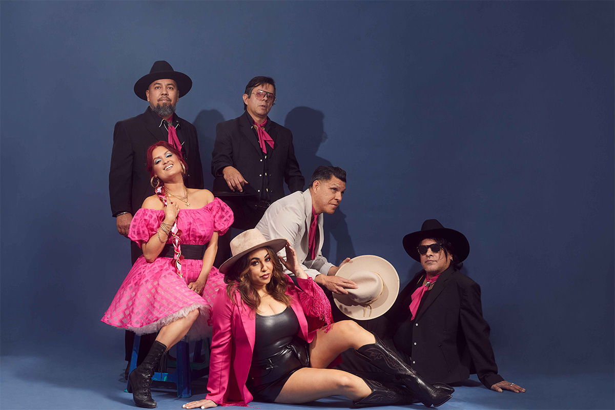 Portrait of the six members of Las Cafeteras, wearing Mexican attire but with pink and black motifs. Two women and four men are part of the band