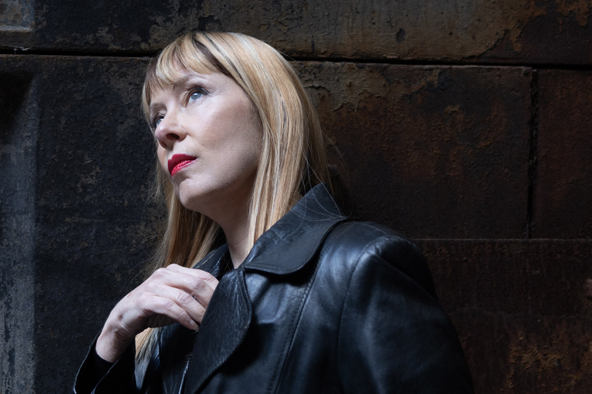 Photo portrait of Suzanne Vega, a woman with blonde hair in a leather jacket looking away from the camera