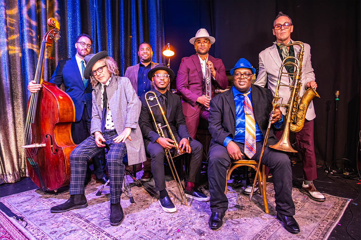 Photo portrait of a jazz band with seven members, all holding their instruments and looking toward the camera. Six of the seven members are wearing glasses.