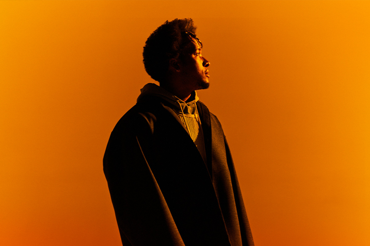 Photo portrait of Cautious Clay, a man with frizzy hair wearing multiple jackets, bathed in orange light