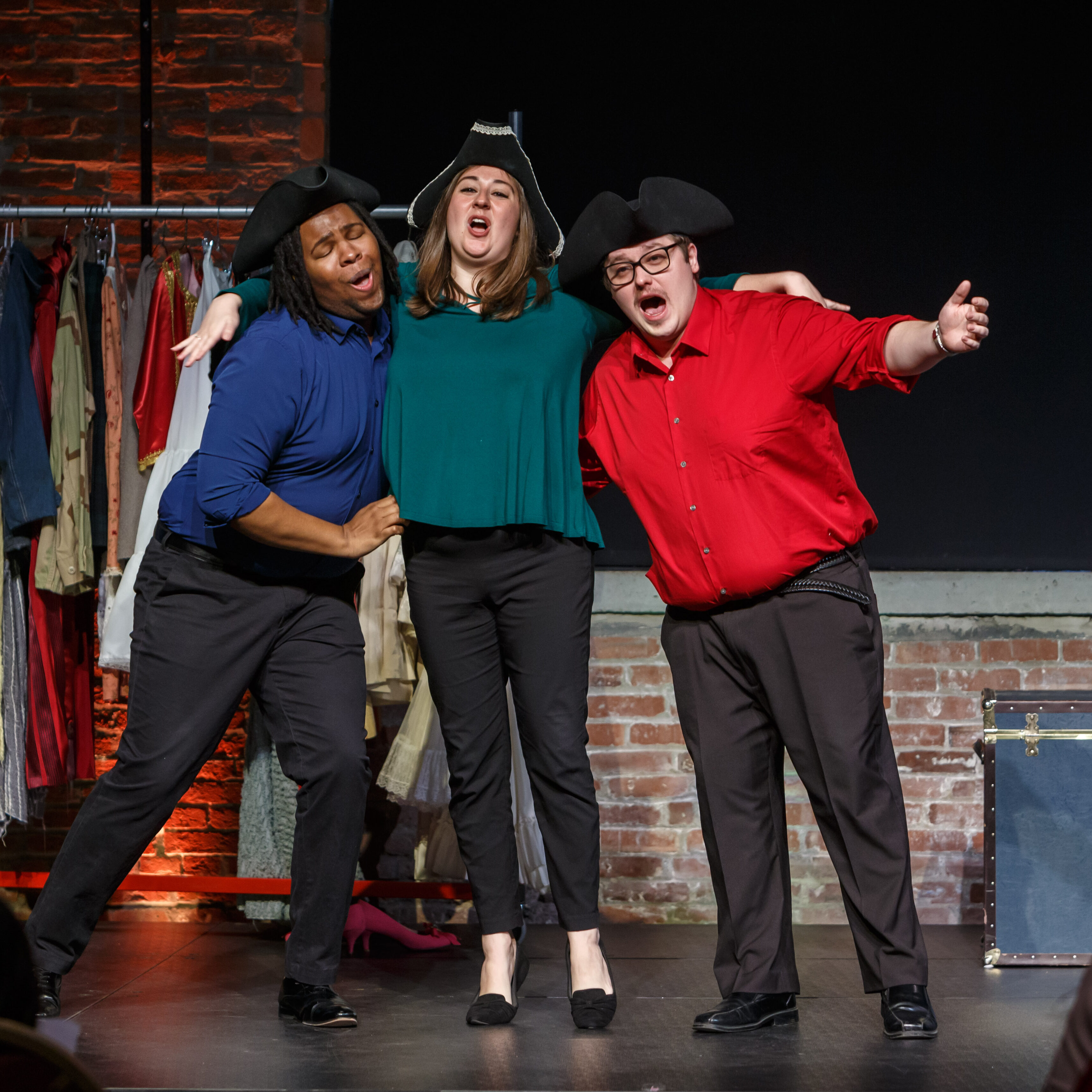 Three Pittsburgh Opera singers, wearing colorful shirts and tri-corner hats, sing shoulder to shoulder on a stage