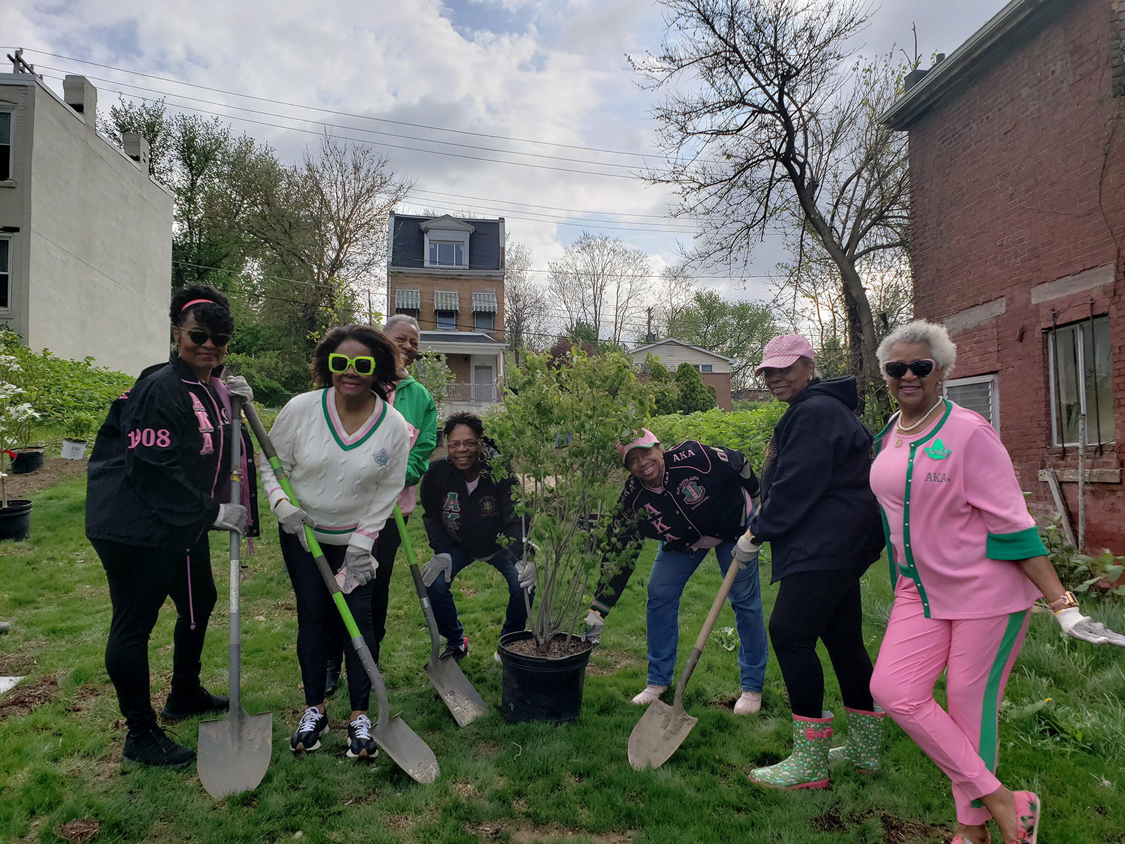 A group of African American women standing around a potted tree with shovels in a green community garden in Pittsburgh's Hill District. They are wearing the pink and green colors of the Alpha Kappa Alpha Sorority.