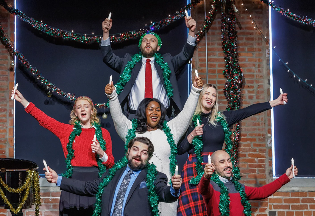 A photo of young Pittsburgh Opera singers in festive Christmas wear, wrapped in garland and holding fake candles on a stage