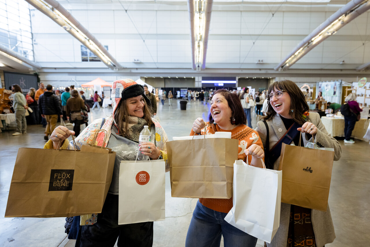 Three women holding bags of purchases at a previous year's craft market at the convention center.